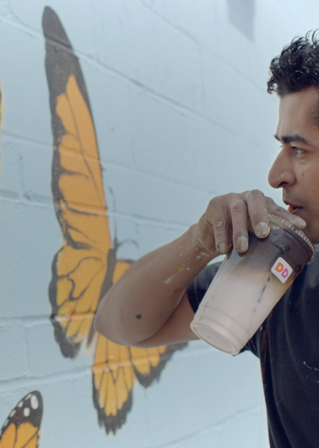 A man drinking a Dunkin' Donuts beverage, next to a mural.