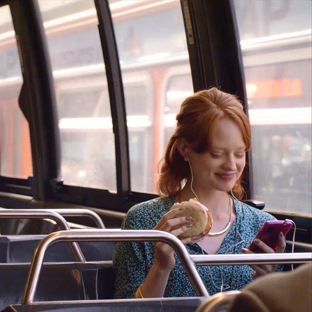A women riding a bus, while smiling and eating a Dunkin' Donuts breakfast sandwich.