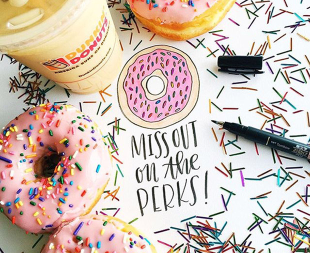 Two strawberry frosted sprinkled donuts and a Dunkin’ Donuts iced coffee on a table with colored confetti.